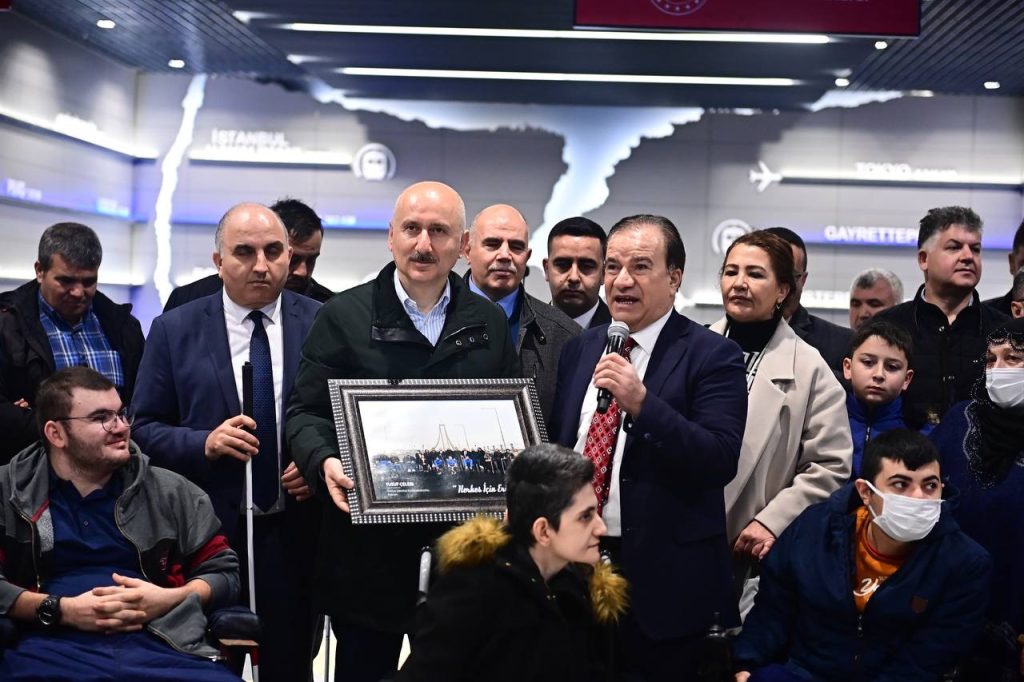 Türkiye Sakatlar Konfederasyonu Başkanı Yusuf Çelebi, Ulaştırma ve Altyapı Bakanı Adil Karaismailoğlu'na bir tablo hediye ediyor ve o sırada mikrofon elinde konuşma yapıyor. Yaklaşık 30 kişi yan yana fotoğrafta görünüyor.