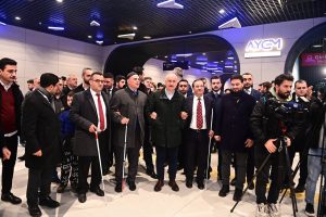 Ulaştırma ve Altyapı Bakanı Adil Karaismailoğlu'nun koluna Türkiye Sakatlar Konfederasyonu Başkanı Yusuf Çelebi ve diğer engelli bireyler girmişler, yan yana duruyorlar.